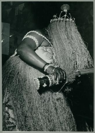 A bare-chested figure wears thick fiber costume and metal collar, standing with hand extended in ritual posture, Bahia Brazil 1962.