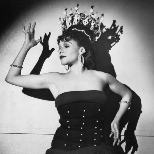 Black-and-white studio portrait of a woman in strapless black dress, long gloves, and ornate crown, right arm raised, gazing upward.