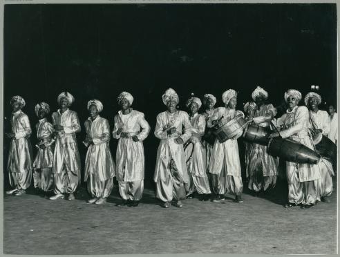 Twenty dancers in white costumes with head wraps stand in a loose line against a dark background, arms in varied positions.