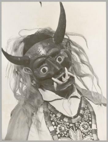 Black and white photograph of a dark carved wooden mask with curved horns, wide grin, staring eyes, and long gray fiber hair attached below the chin.