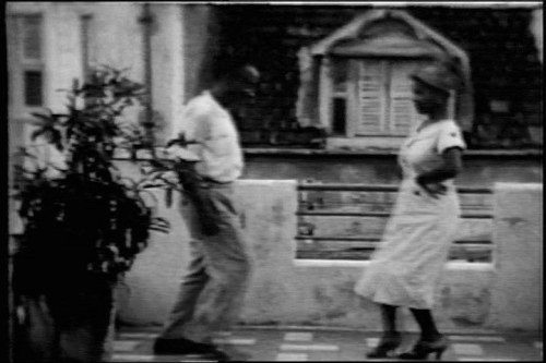 Two dancers in light-colored clothes dance outdoors in a residential yard in Jamaica, with wooden buildings visible behind them.