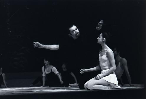 Black and white photograph of a shirtless male dancer seated and gesturing toward a standing female dancer in white, surrounded by an ensemble on a dark stage.