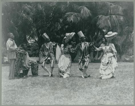 Six women in white dresses and wide hats stand beneath tall palm trees in a tropical outdoor setting, holding fans or similar objects.