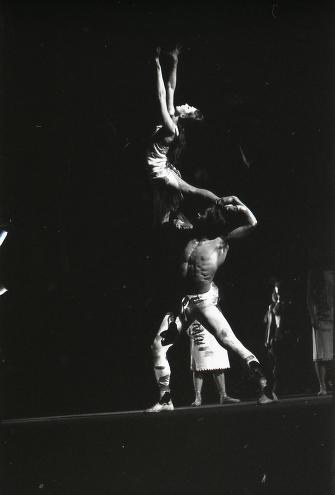 Male dancer in wide stance supports inverted female dancer in extended arch against dark stage backdrop, strong backlighting.