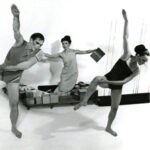 Four modern dancers suspended in mid-leap against white background in coordinated but separate positions, arms extended, bodies arched.