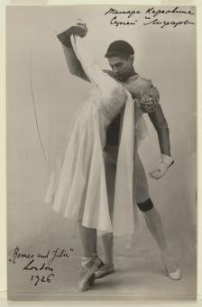 Two ballet dancers in studio costume: man in dark tunic and tights with one arm raised, woman in pale shift dress with draped shawl, both en pointe.
