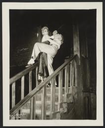 Black-and-white photograph: a male dancer in white tie and tails lifts a female dancer in a white tutu high overhead on a staircase.