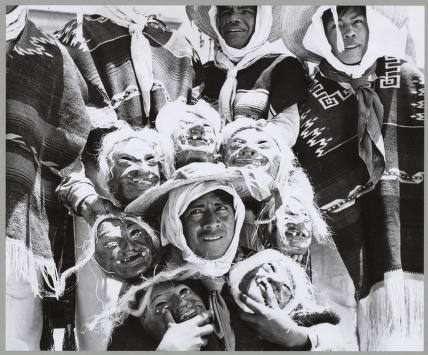 Group of people wearing carved masks and woven regalia, tightly clustered and facing the camera in a black-and-white photograph.