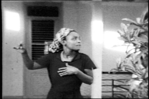 Black-and-white photograph of a woman in a dark shirt dancing alone indoors, one arm raised, captured mid-movement in what appears to be fieldwork