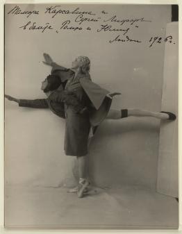 Ballerina in dark coat holds an extreme arabesque penché while male dancer in dark clothes frames her line in white studio.