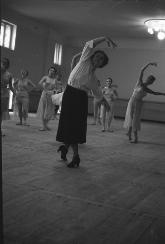 Black-and-white photograph of a woman in ballet practice clothes demonstrating an overhead arm position to a group of young ballet students in a studio.