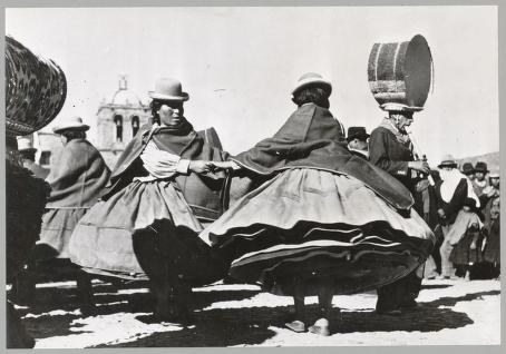 Two dancers in full traditional Andean skirts spin outdoors, skirts billowing, with spectators and equipment visible in background.