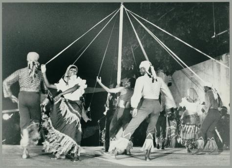 Four dancers spiral around a central maypole with radiating ropes, their bodies leaning inward as their skirts and clothing catch the rotational movement.