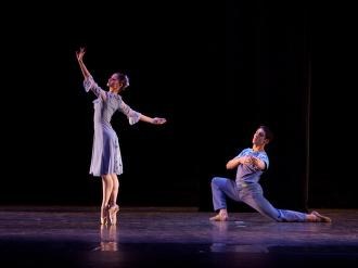 Two ballet dancers on a dark stage: one in a tutu en pointe with raised arm, the other on the floor in a kneeling lunge position.