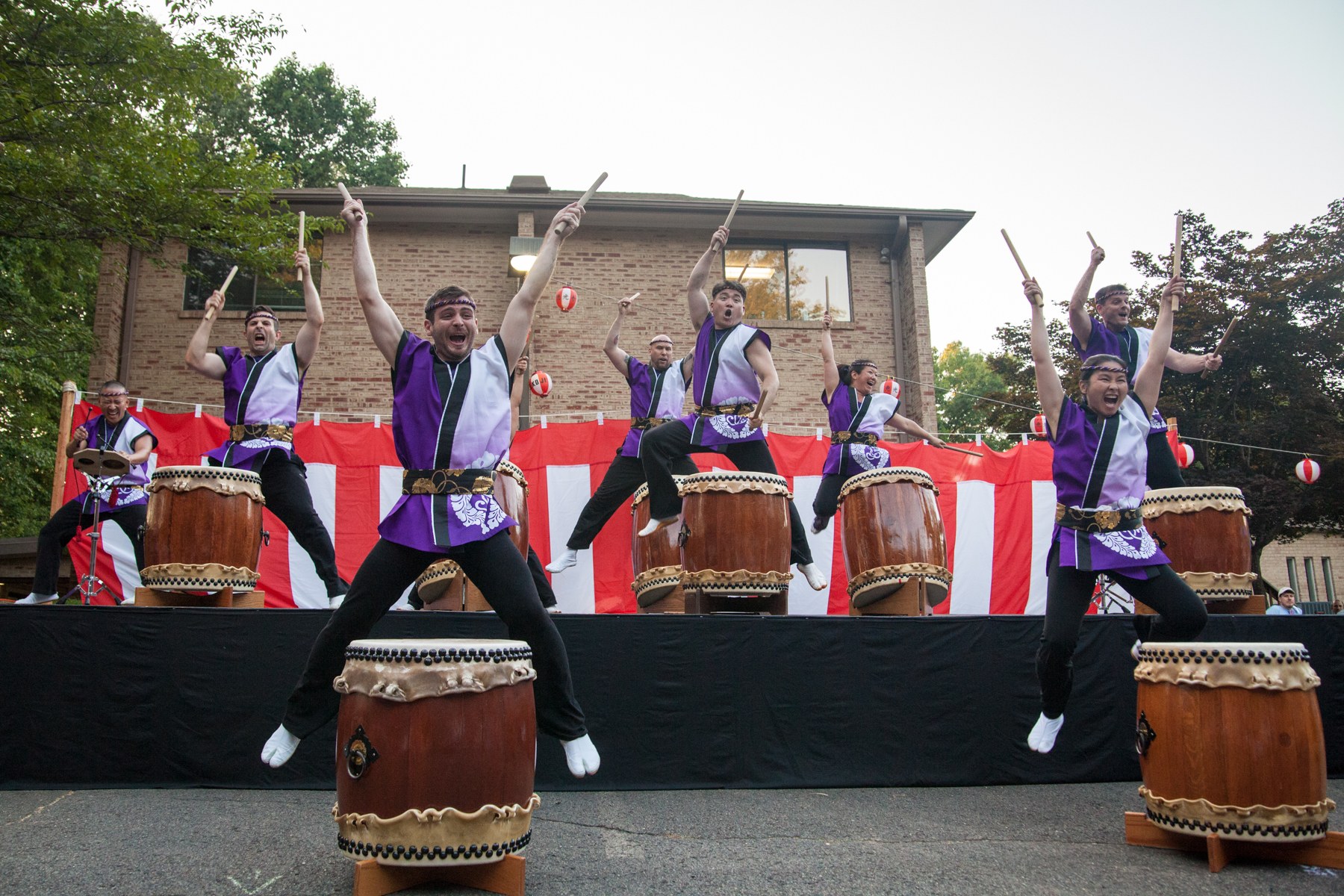 A taiko drum ensemble in purple and white costumes stands on stage with raised drumsticks, performing with open mouths and raised arms.