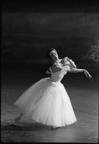 A ballerina in a white tutu and pointe shoes stands on a dark stage, arms raised in a romantic ballet pose.