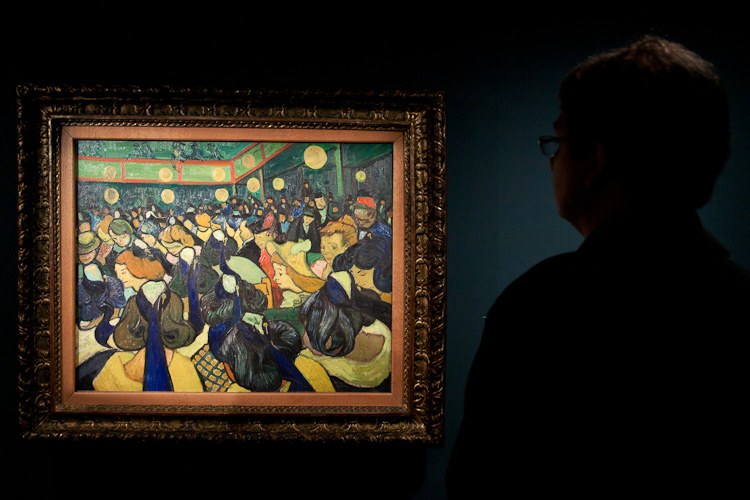Man in silhouette viewing van Gogh's expressionist painting of a dimly lit café interior with figures seated and standing among tables and lamps.