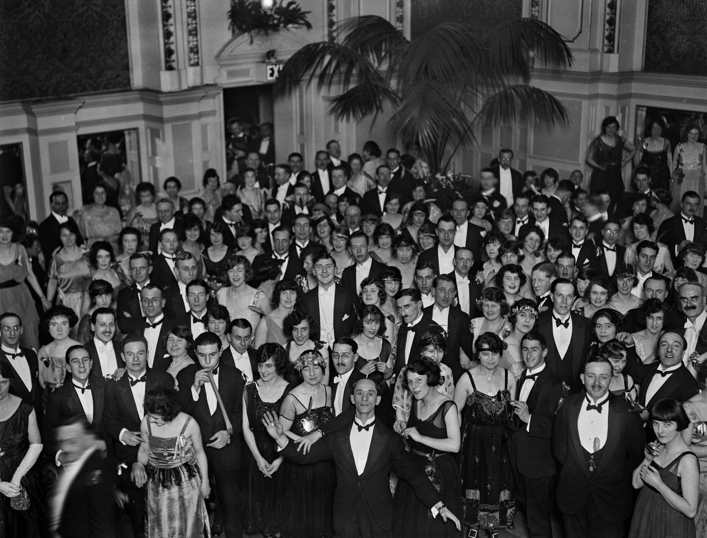 Crowd of about 100 formally dressed men and women at a 1921 ballroom dancing competition, with a man in a light jacket standing centrally among darker-suited