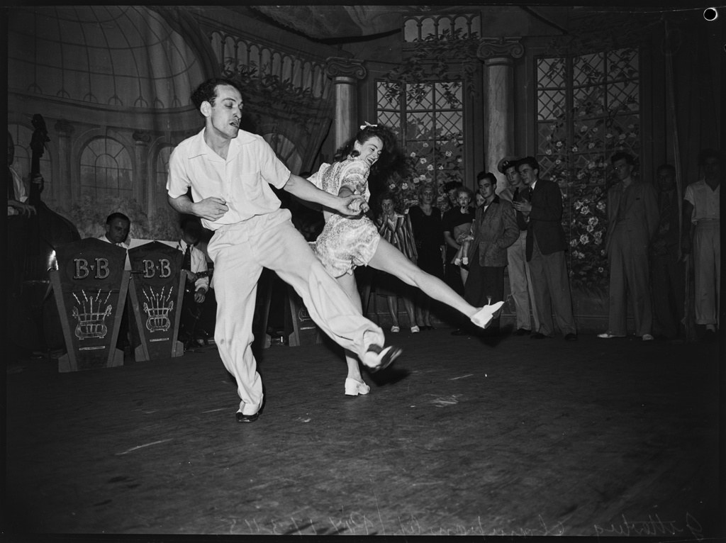 Male dancer in white suit lifts female partner horizontally above a ballroom floor while formally dressed spectators watch, 1948.