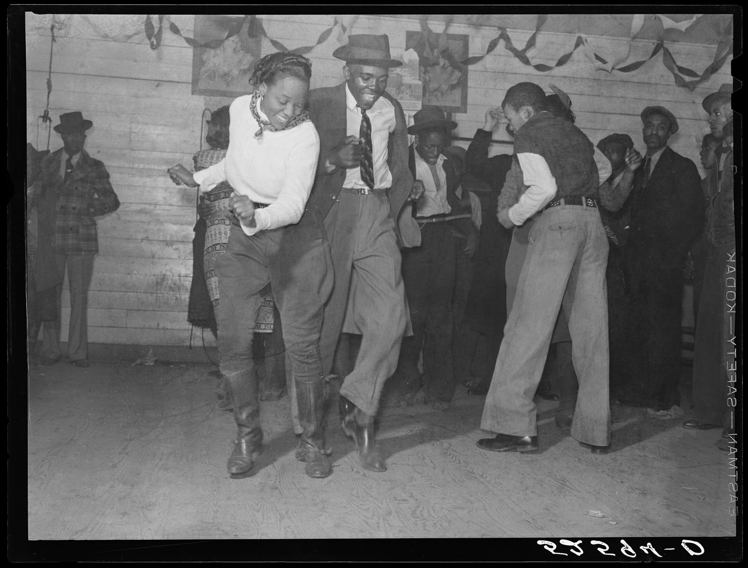 Three couples dance in close hold on a wooden floor in a crowded juke joint, the nearest pair in center focus, wearing 1930s casual clothing.