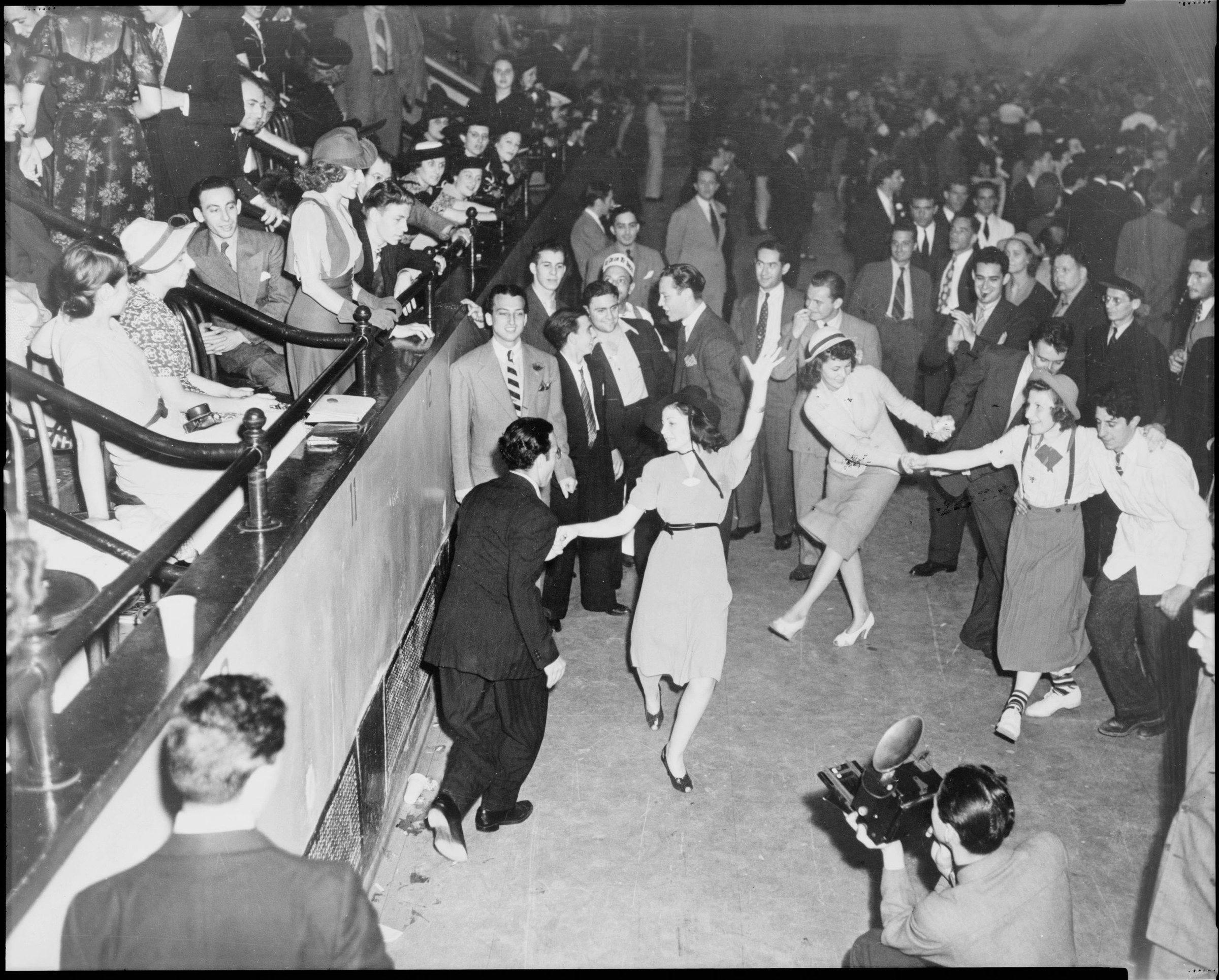 Couples jitterbug dancing in center of crowded indoor venue surrounded by spectators in 1930s dress, with press photographer in foreground, 1938.