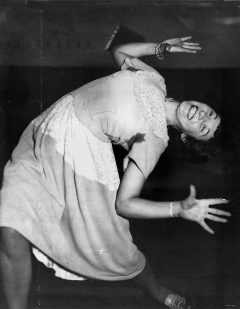 A woman in a light dress arches backward mid-dance, arms spread, in a dynamic jitterbug pose.