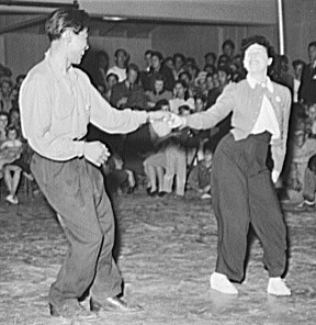 Two couples perform jitterbug in a rigid partnered frame at a community dance competition, watched by spectators seated in the background.
