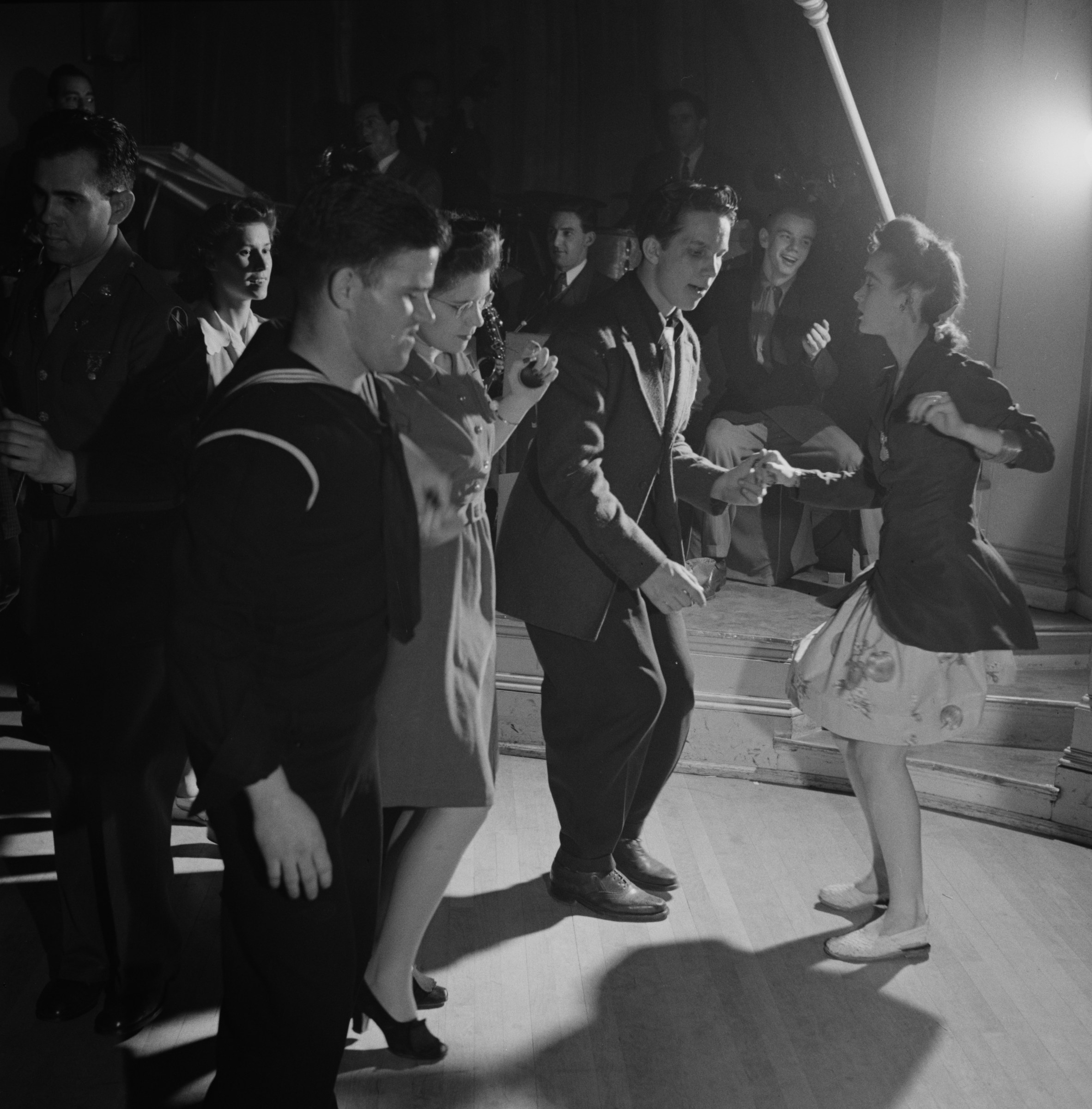 Crowded dance floor with couples in swing position; foreground woman's skirt flares mid-turn under bright spotlight, 1943.