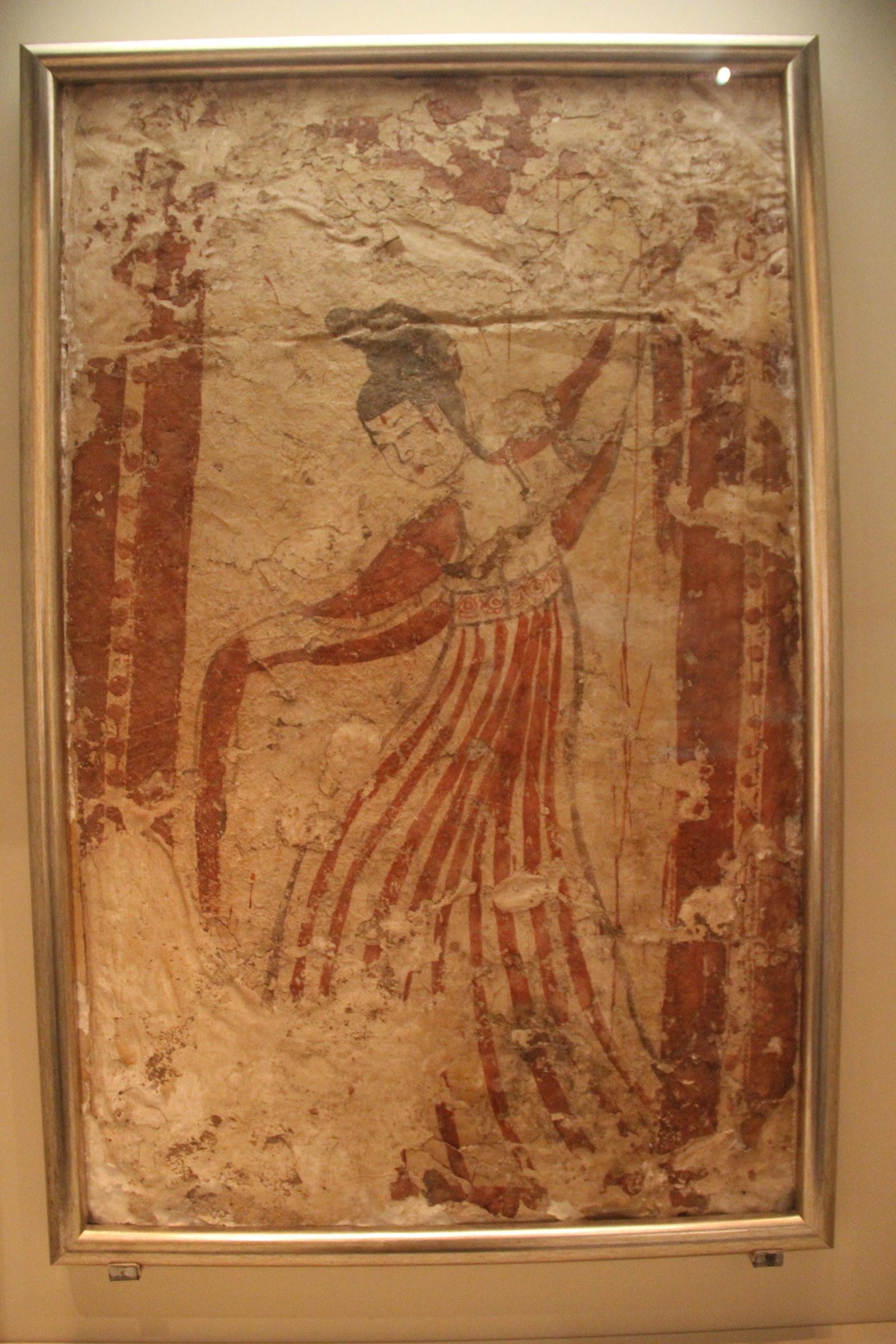 Ancient Tang Dynasty fresco showing a woman in pleated skirt and ornate belt holding a staff, torso twisted and arm extended, in an interior columned space.