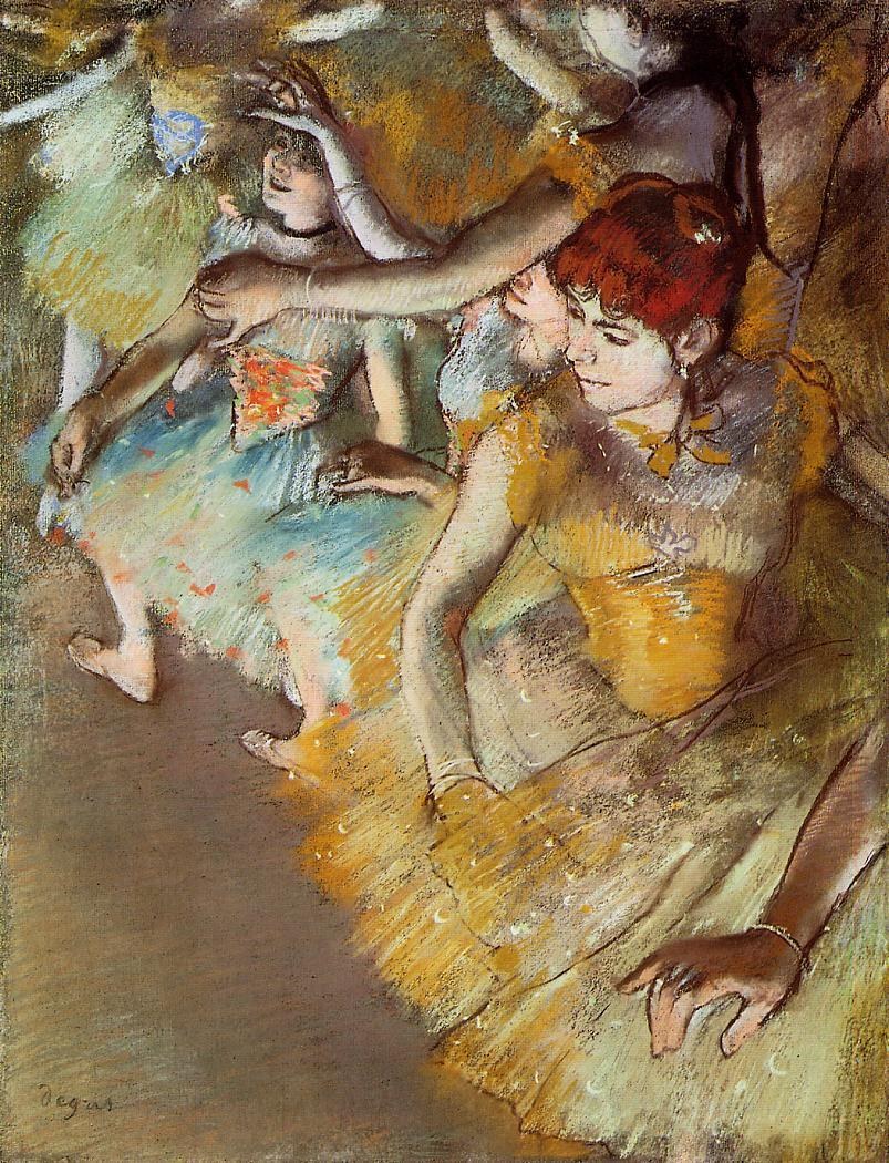 Three young female ballet dancers in pastel tutus rest and stretch on a dimly lit stage, arms and legs extended in classical positions.