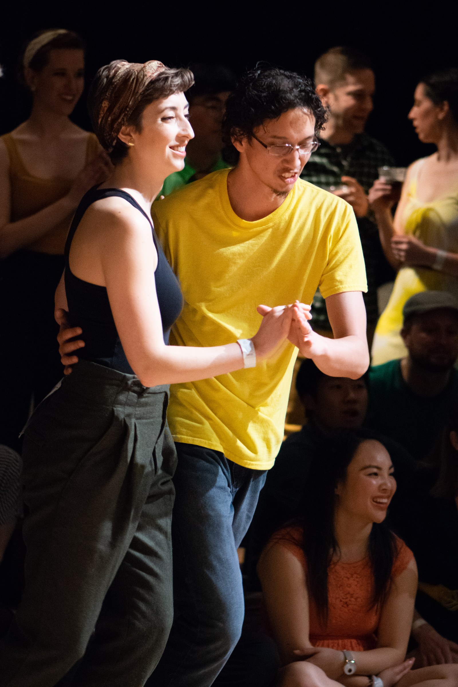 Man in yellow shirt and woman in black top smile at each other while dancing, surrounded by seated and standing spectators in a darkened venue.