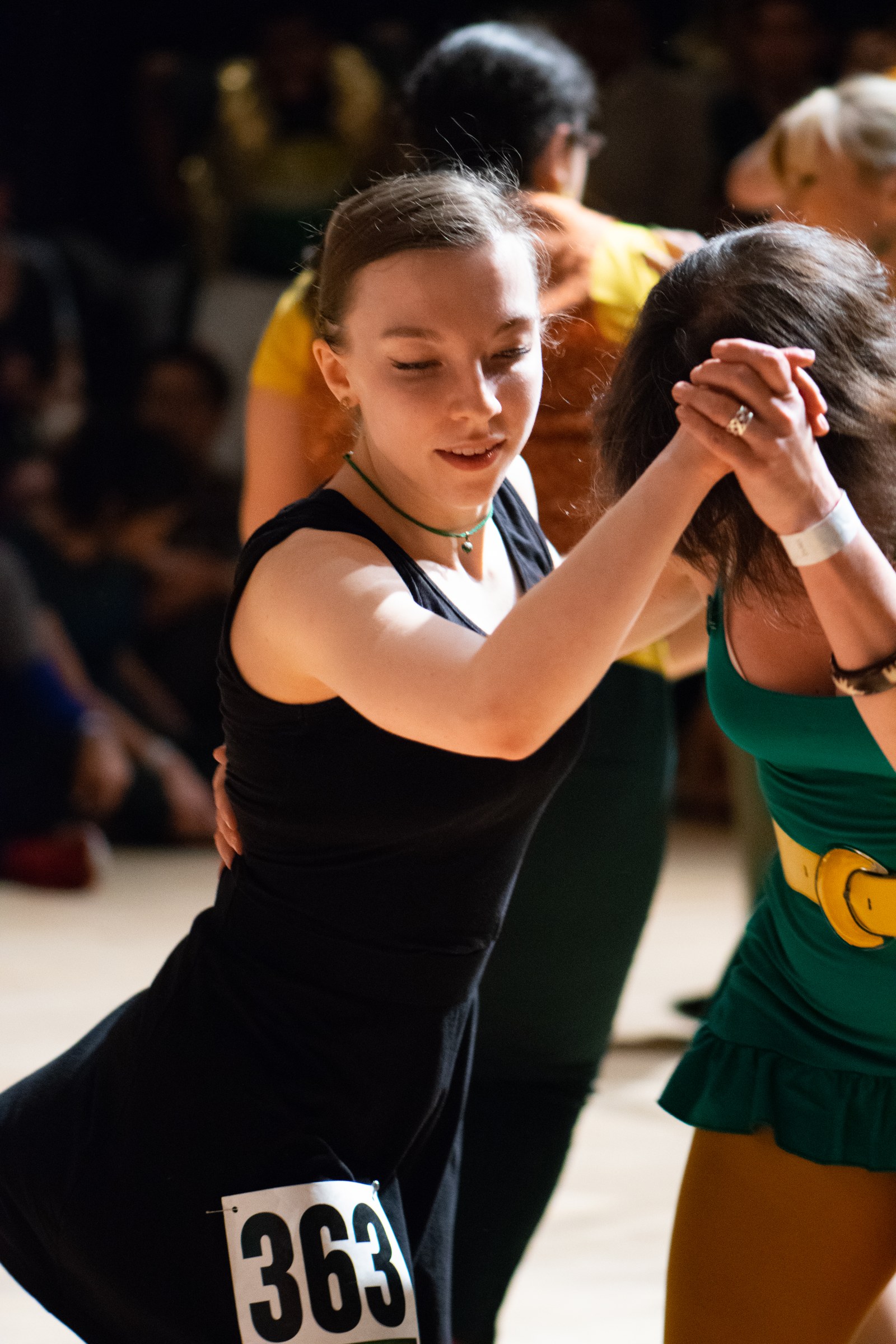 Young female dancer in black competition dress mid-movement at an indoor swing dance competition, wearing number 363.