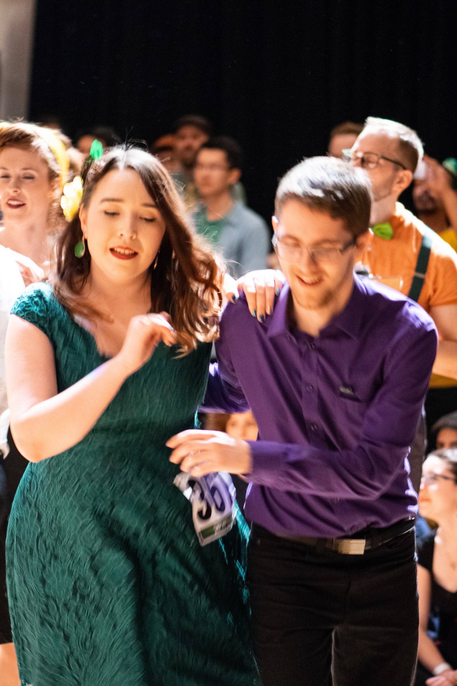A couple dances Lindy Hop at a 2018 swing competition. The woman wears a teal dress with a yellow flower clip; the man wears a purple shirt.