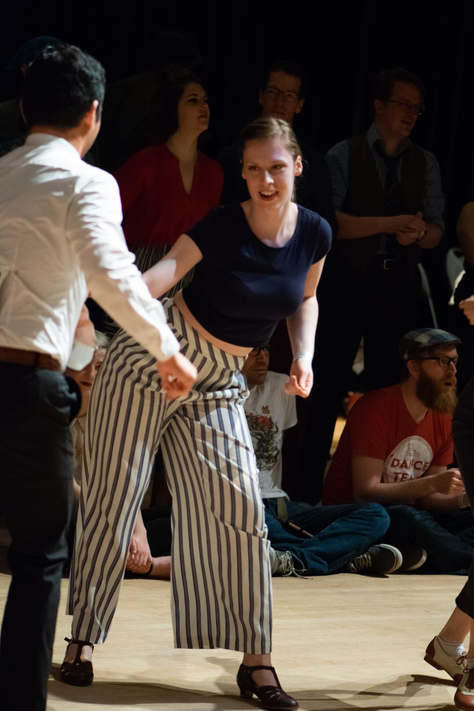 Lindy Hop dancer in navy shirt and striped trousers performs with a partner in white shirt on a wooden floor, surrounded by seated and standing spectators in