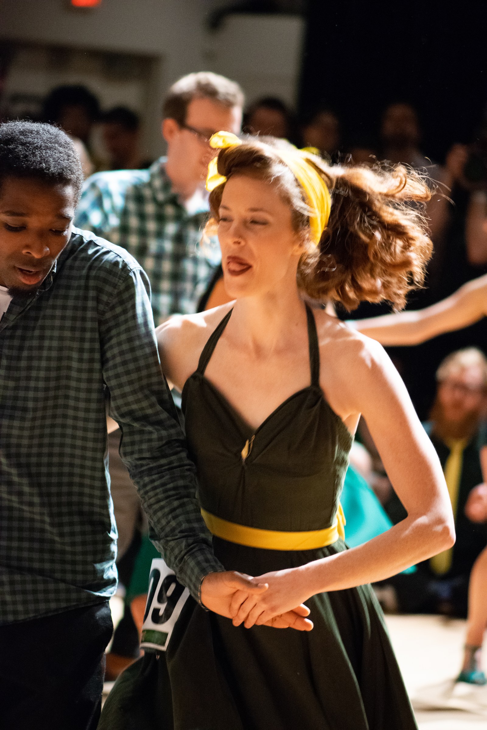 Woman in black dress with yellow belt and man in checked shirt dance hand-in-hand at a Lindy Hop competition, both upright and engaged.