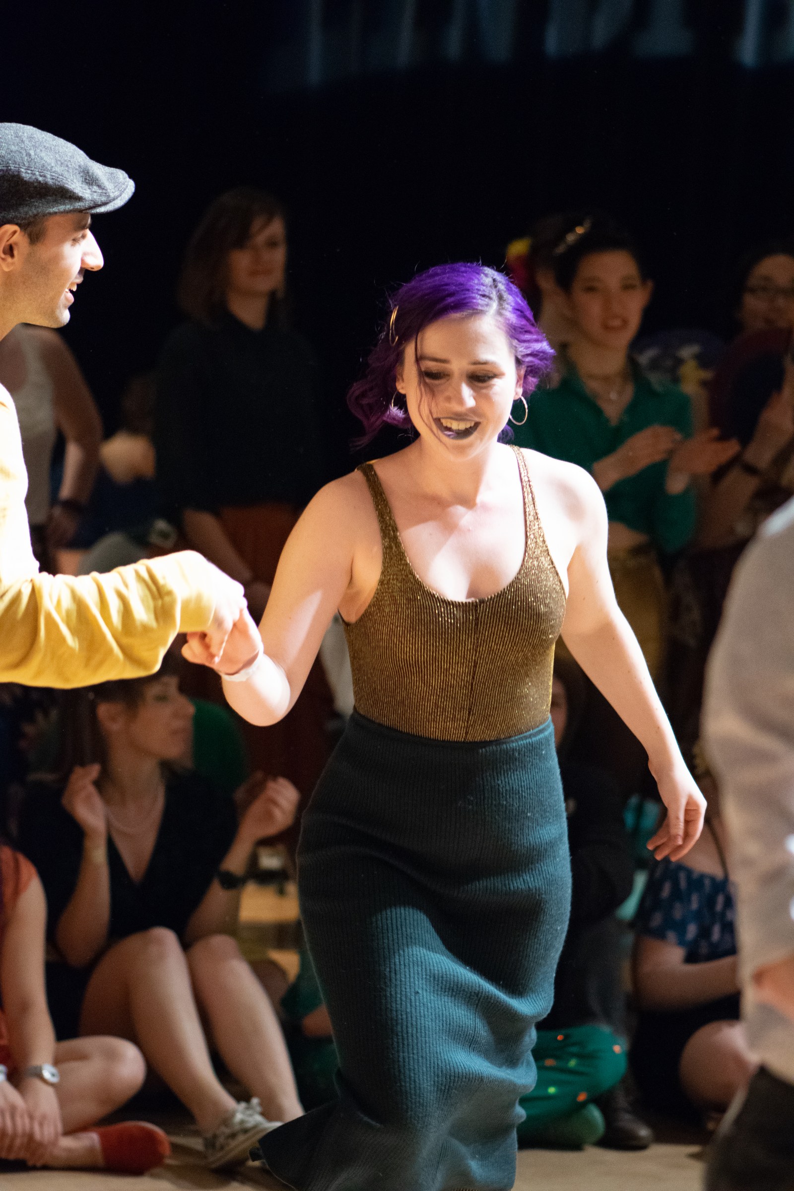 Woman with purple hair and man in flat cap dance in closed position at a swing competition, surrounded by spectators