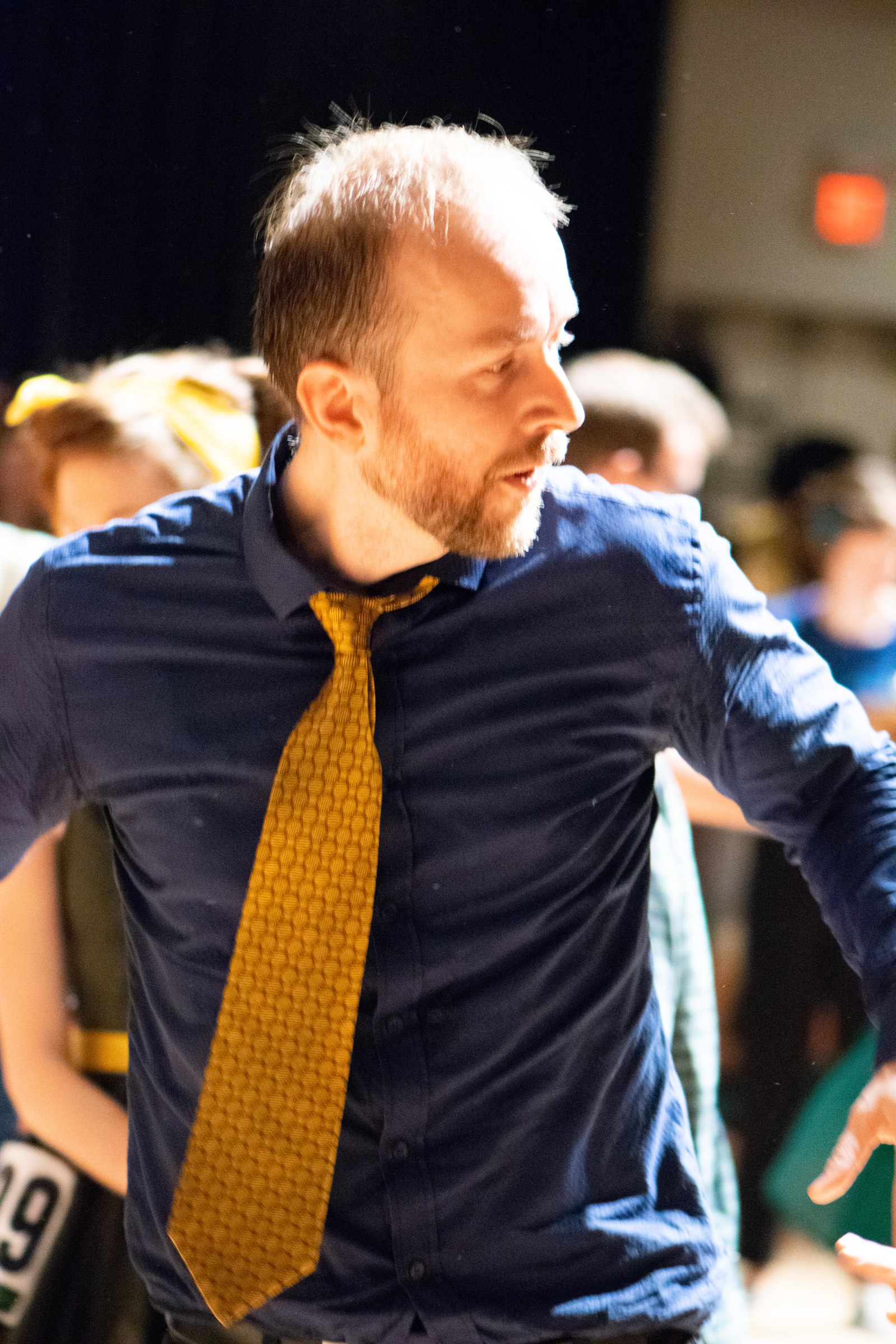Man in navy shirt and gold tie leans forward mid-dance at a swing competition, mouth open with effort and intensity.