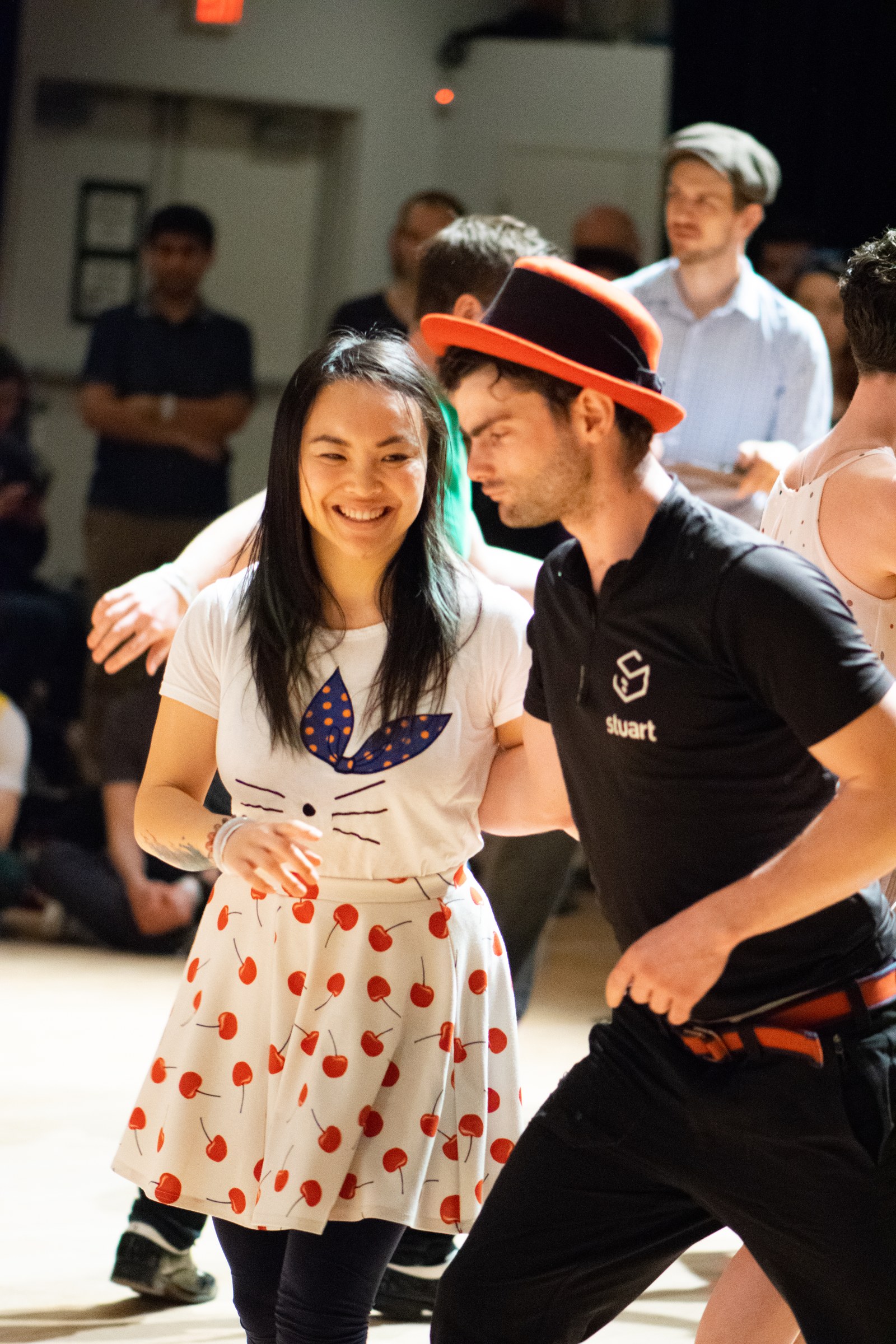 Two smiling dancers in casual modern dress perform Lindy Hop on an indoor dance floor, surrounded by watching spectators at a 2018 swing dance competition.