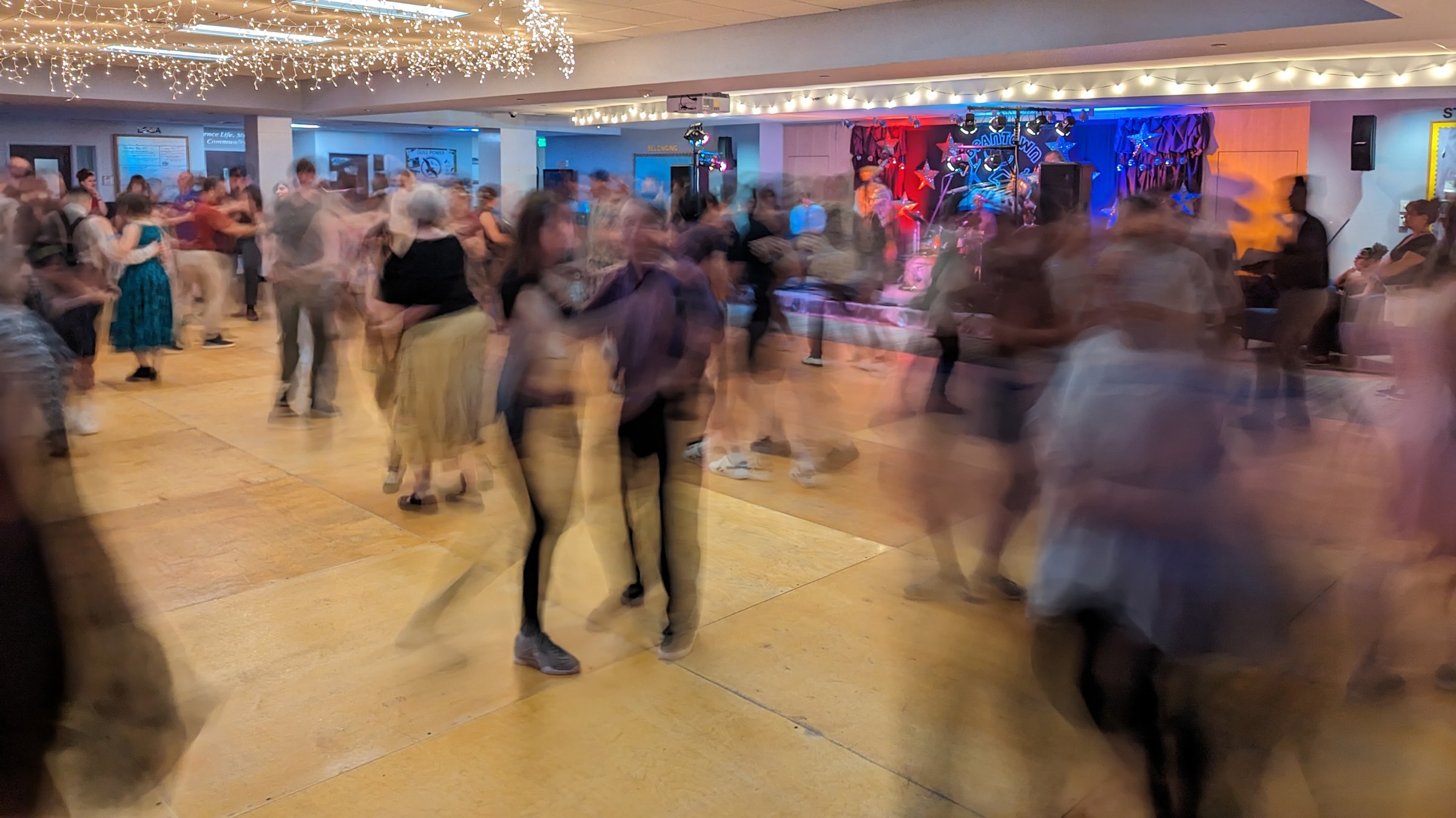 Long-exposure photograph of dancers in blur on a wooden floor under chandeliers and colored lights at an indoor swing dance event.