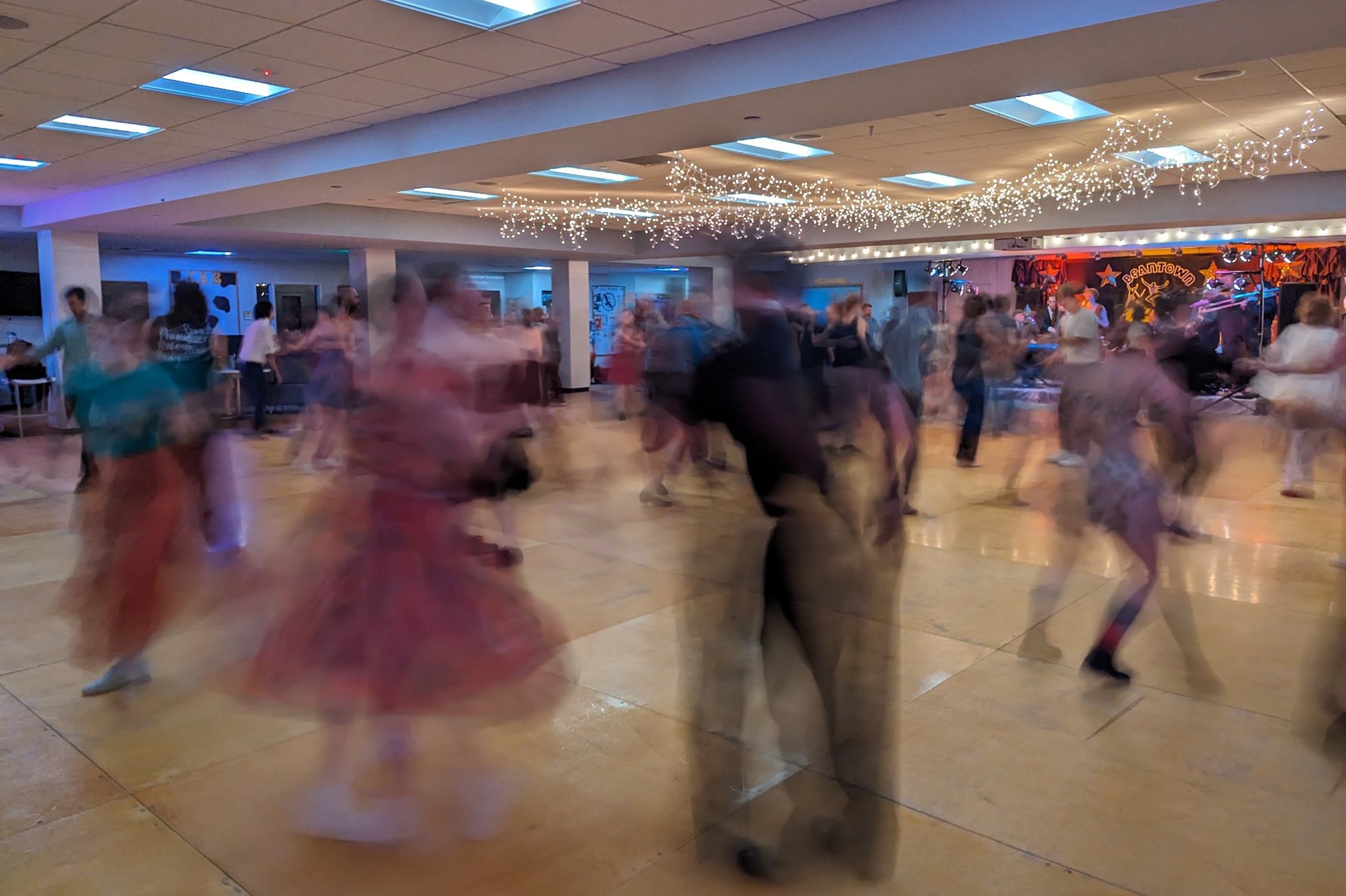 Long-exposure photograph of dozens of Lindy Hop dancers in motion across a polished wooden floor in a brightly lit event space, their blurred and overlapping