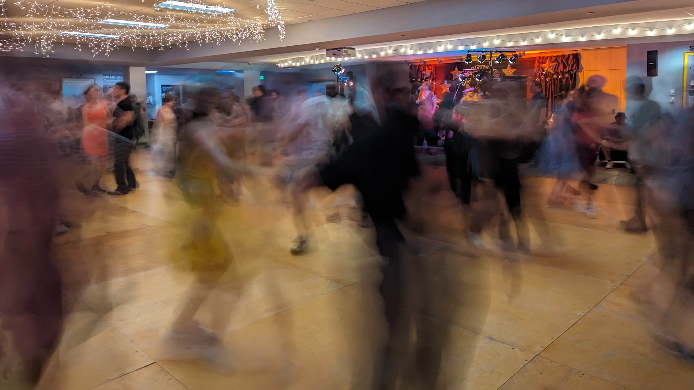 Long-exposure photograph of dozens of blurred dancers moving across a wooden floor in a brightly lit indoor venue.