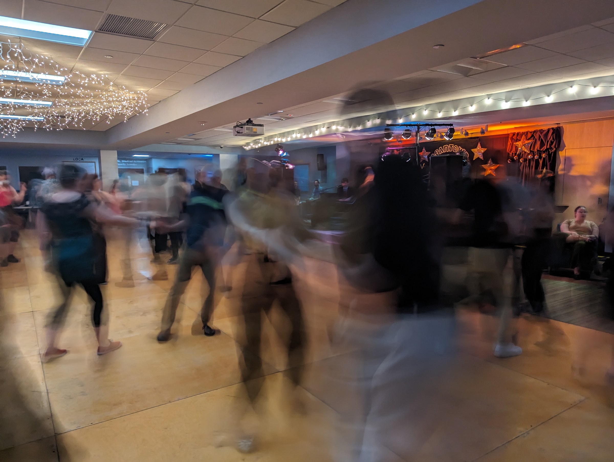 Long-exposure photograph showing dozens of blurred Lindy Hop dancers in closed hold on a wooden ballroom floor, lit in amber and orange, with spectators seated