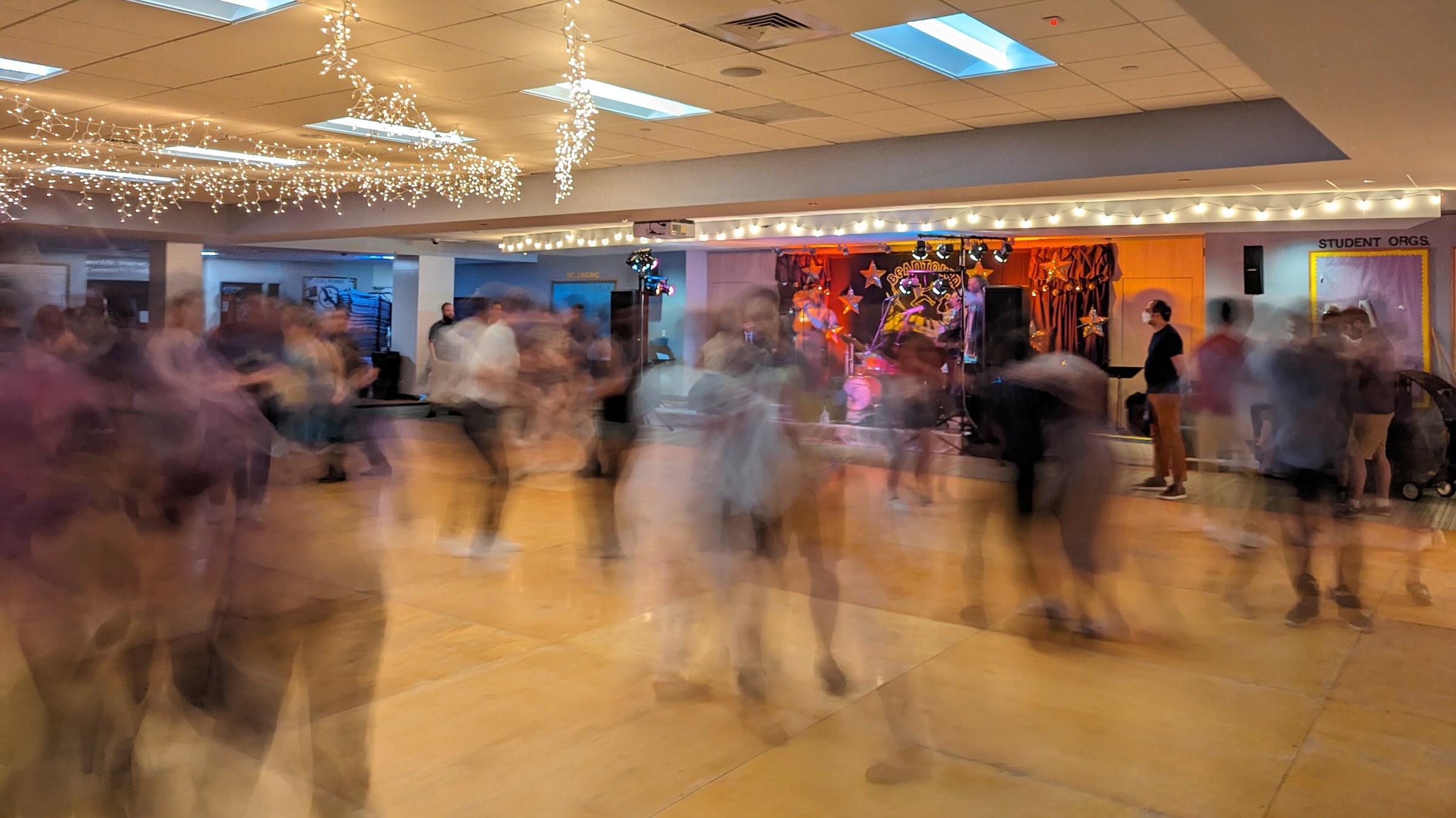 Long-exposure photograph of dozens of Lindy Hop dancers moving together on a wooden floor in a modern hall with warm lighting and pastel walls.