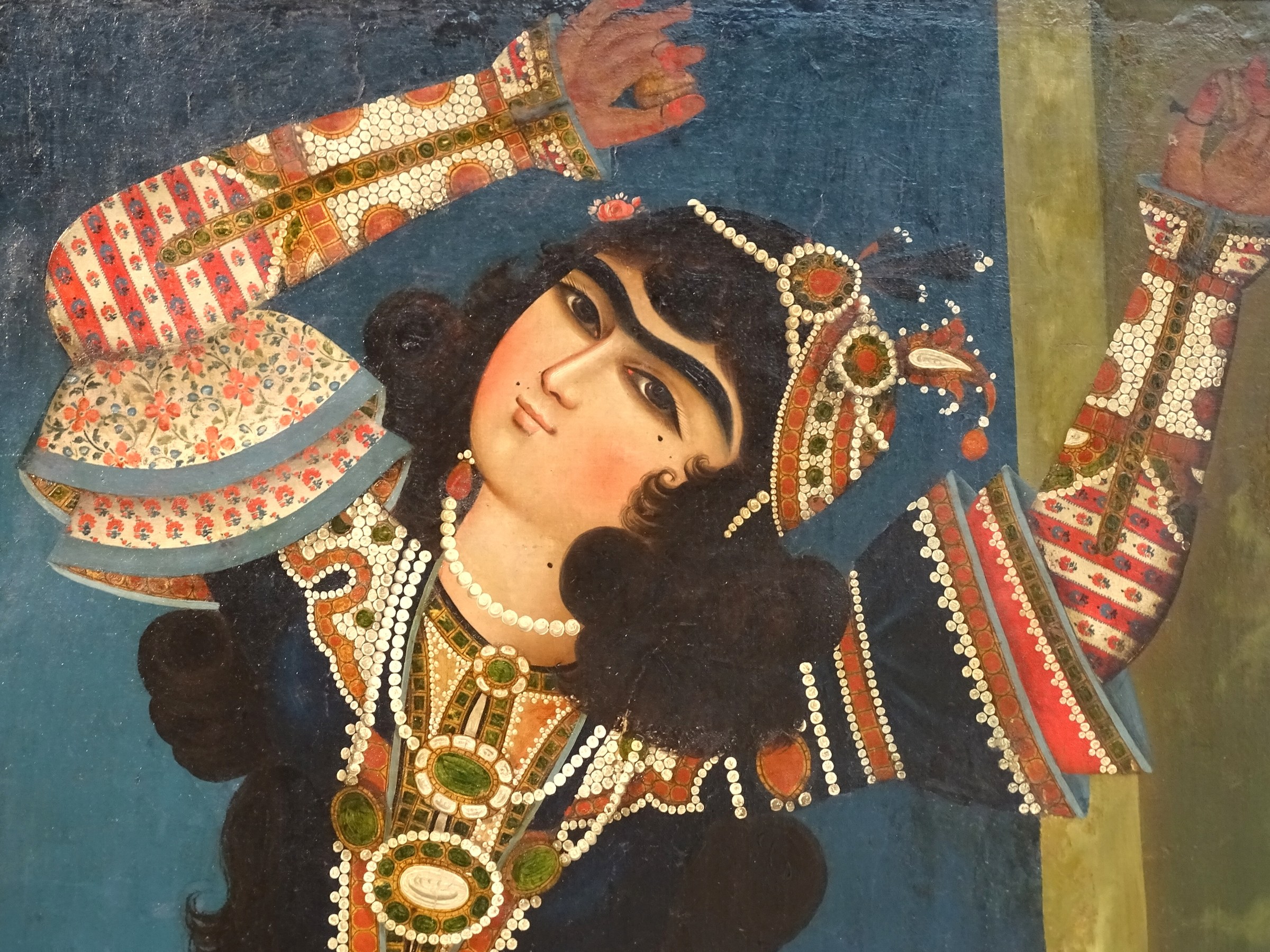 Painted figure of a Persian dancer wearing an ornate jeweled headdress and patterned robes, arms raised in expressive pose, holding a castanet.