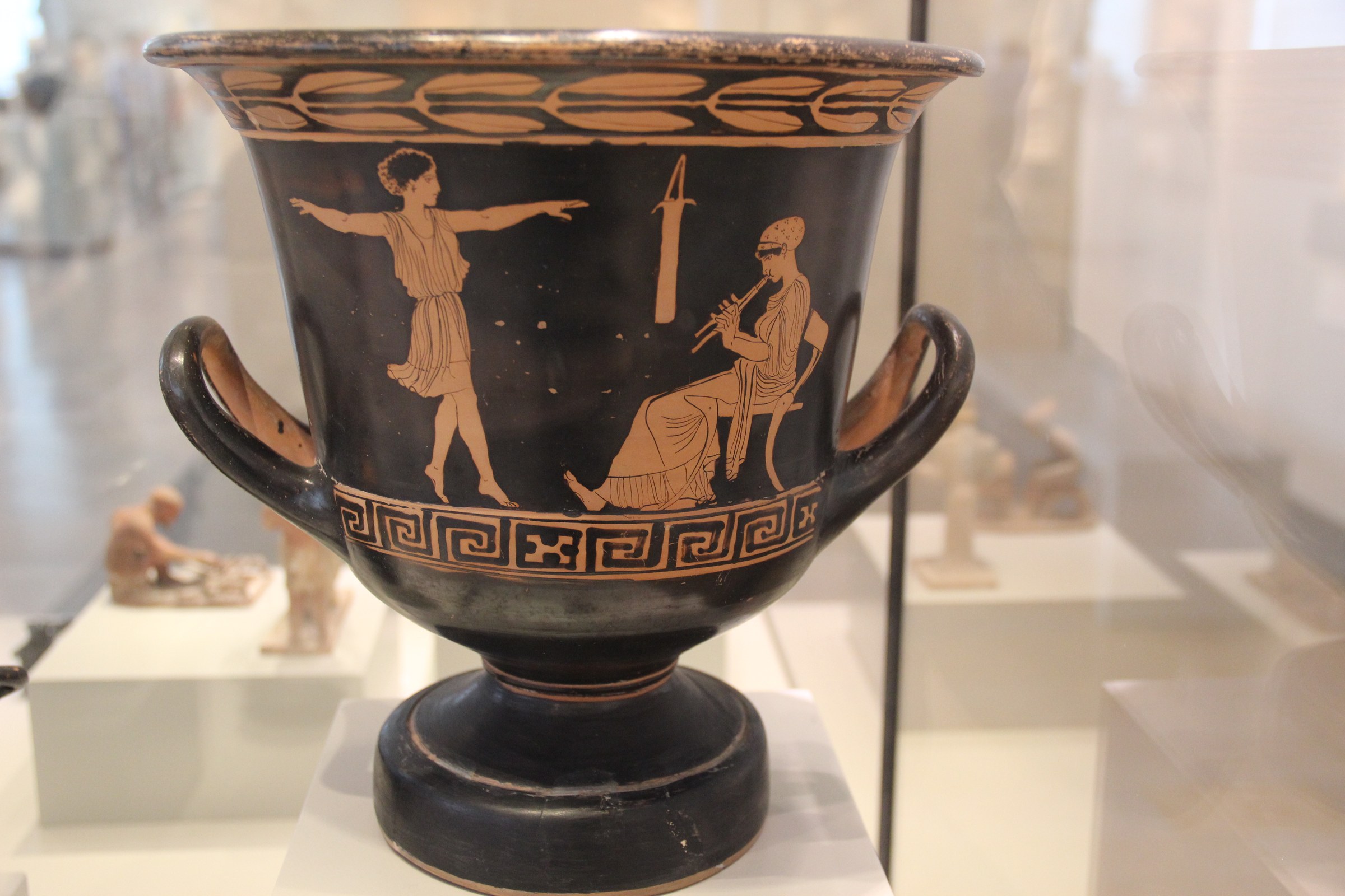 Ancient Greek red-figure calyx crater showing a young bearded man with raised arm facing a seated musician holding a double flute, with an altar between them.