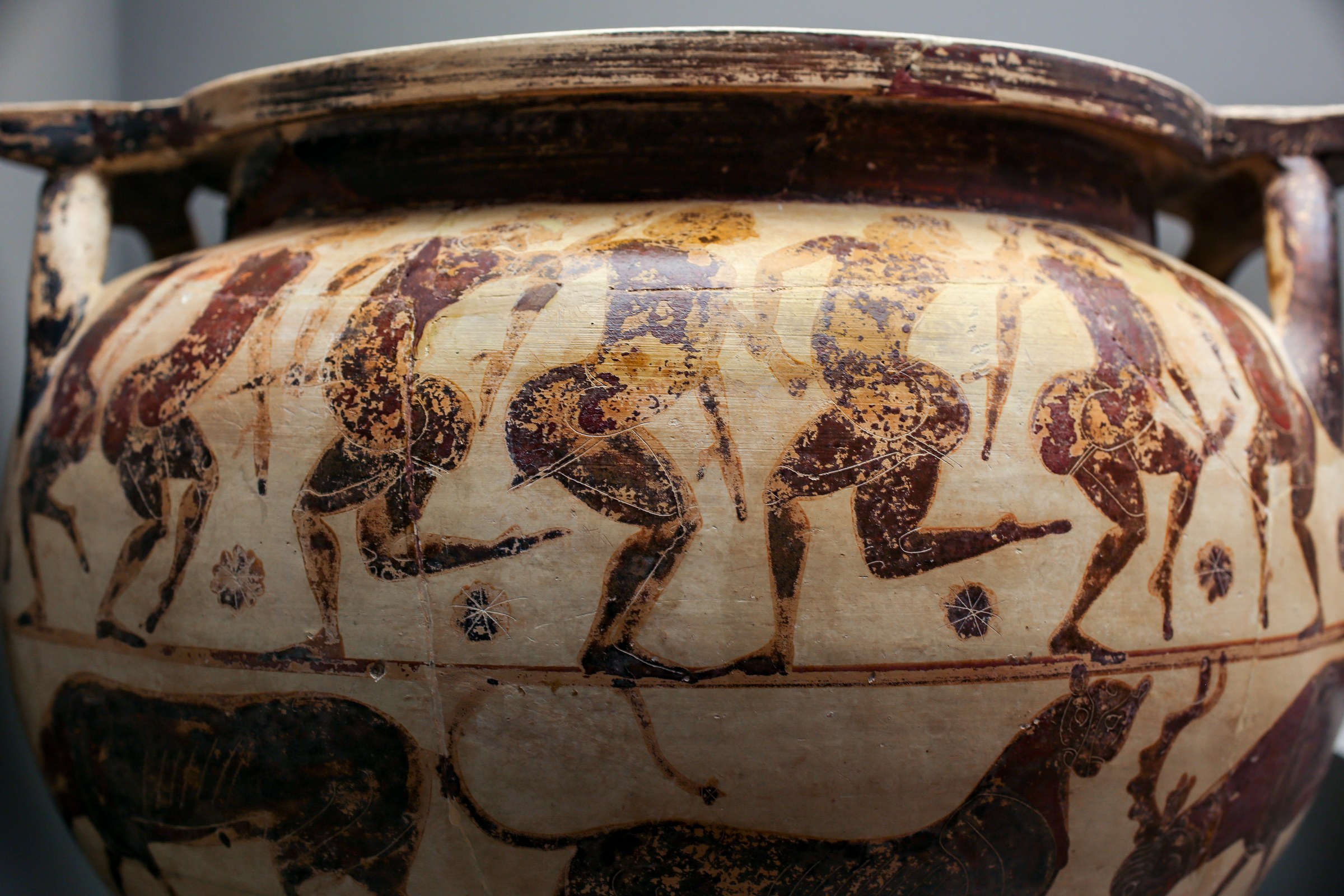 Five padded male figures in profile stand around a ceramic bowl, arms raised in celebratory gestures, separated by floral ornaments.