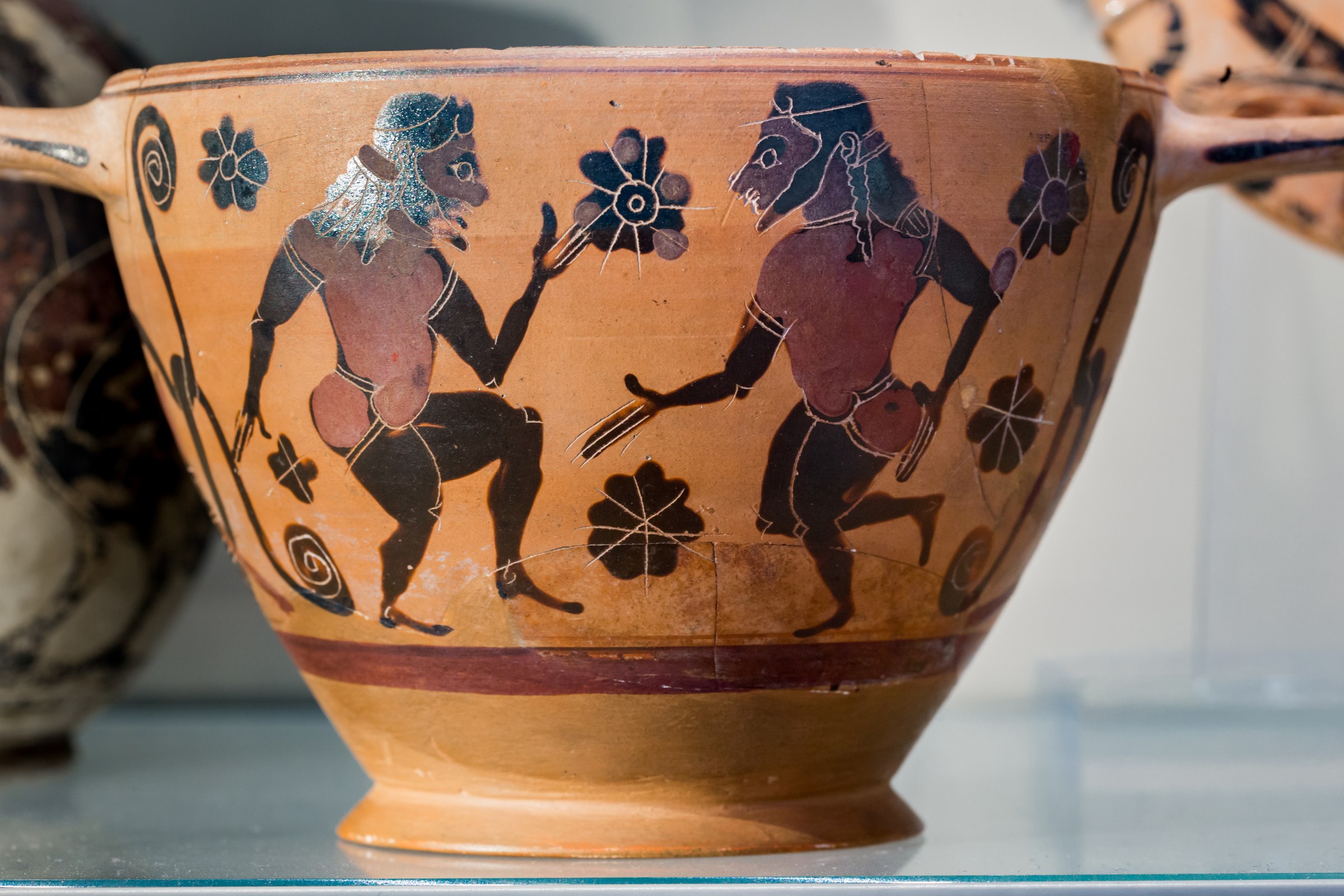 Terracotta cup with two bearded men in grotesquely padded costumes dancing and holding flowering plants.