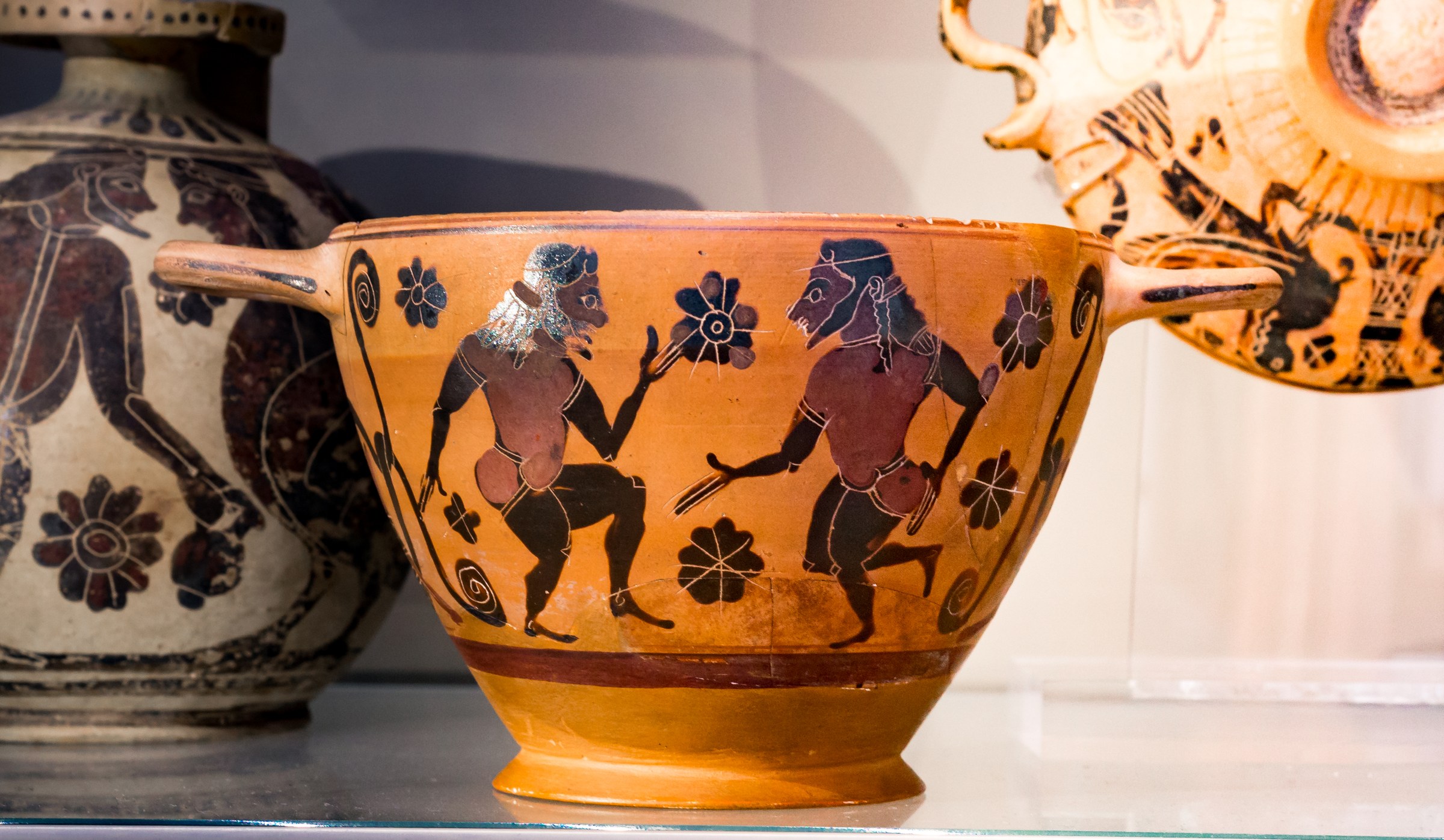 Two bearded men in padded costumes face each other in dynamic poses on an orange terracotta cup, surrounded by small flowers.