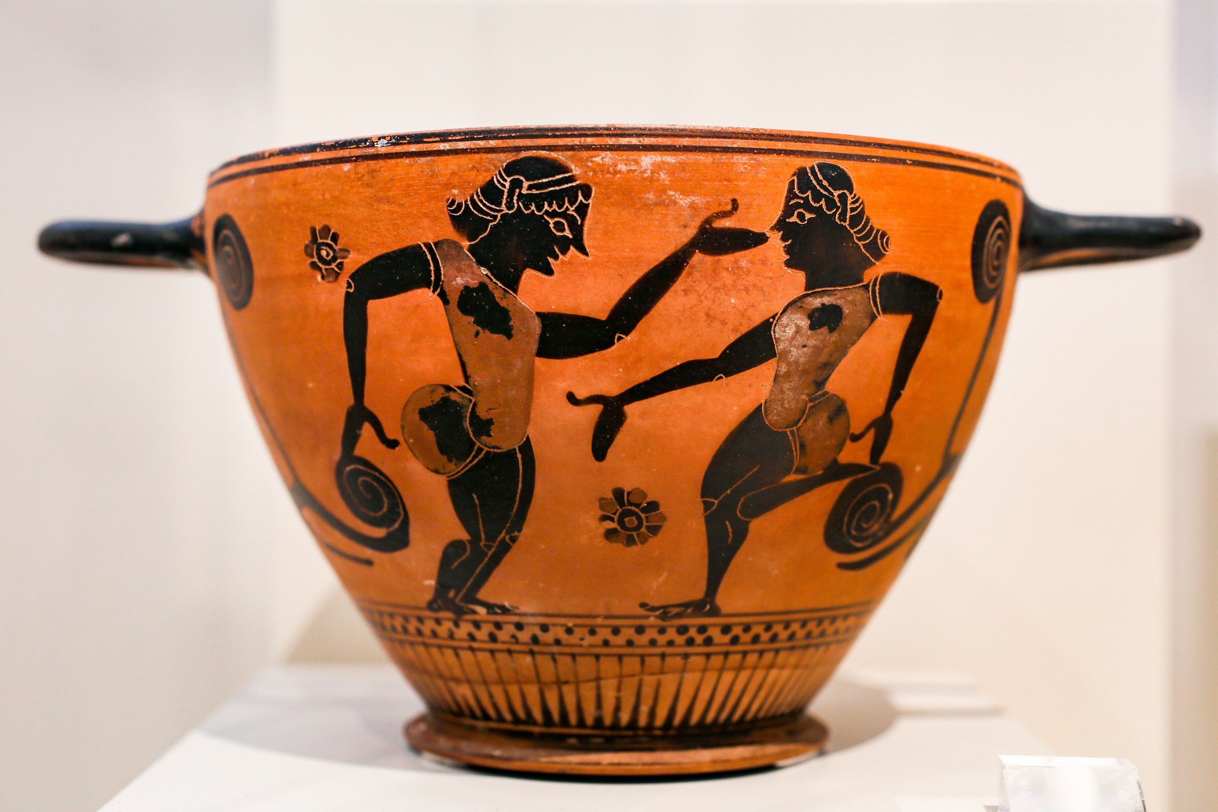 Two bearded men in bulbous padded costumes dance facing each other on an orange terracotta cup, with a pipe player on the reverse.
