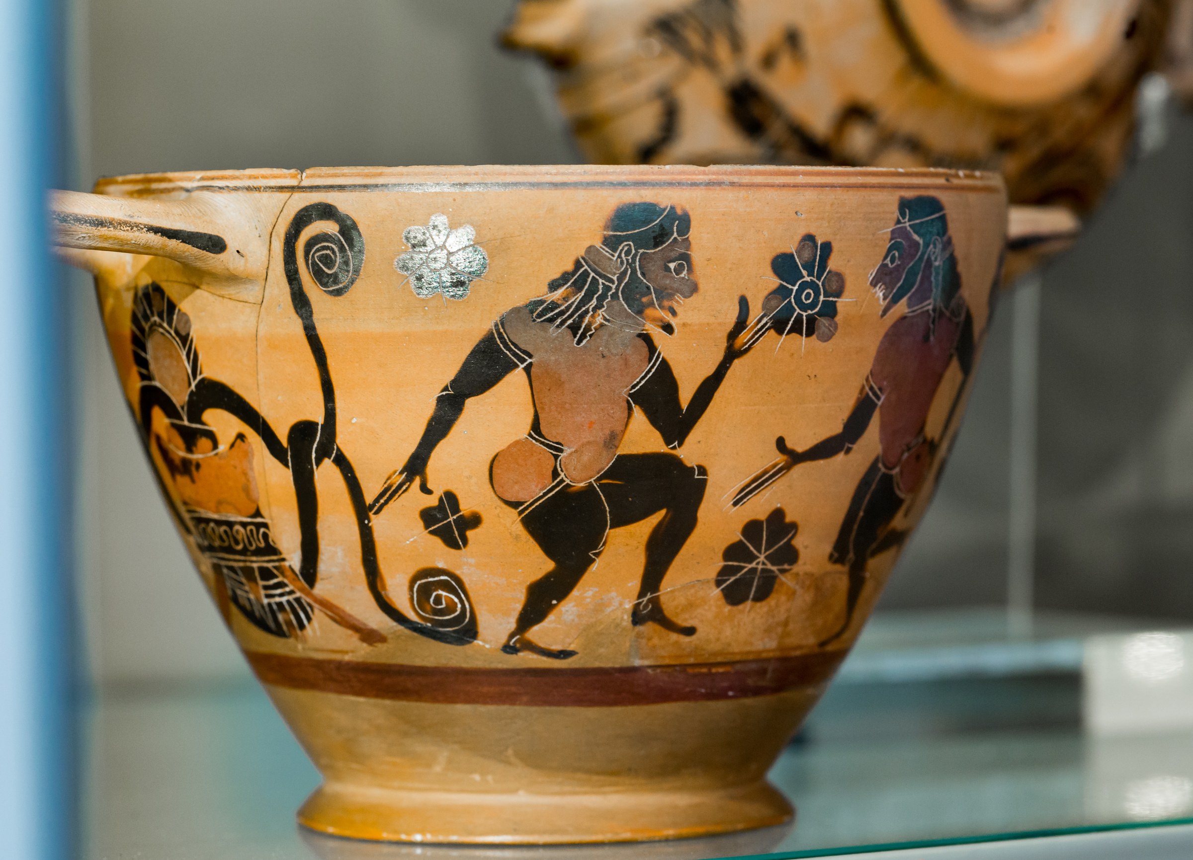 Terracotta drinking cup with black figures: two bearded men in padded costumes with wide-legged stances and raised arms, surrounded by floral motifs.